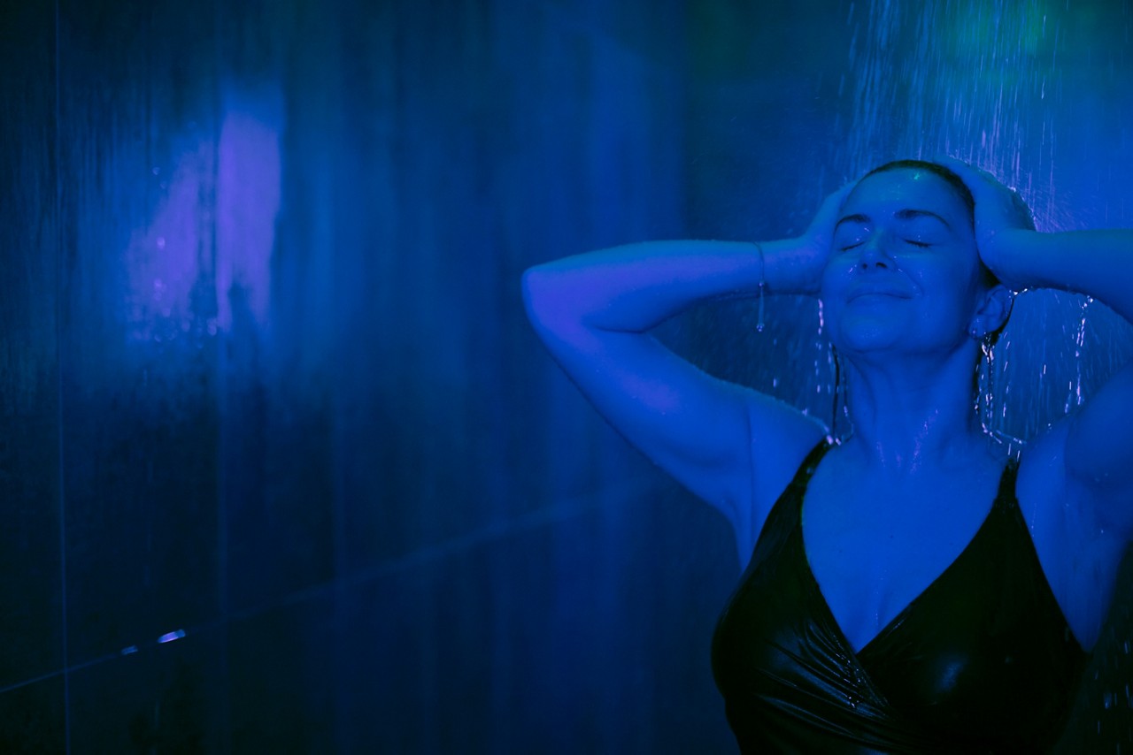 Person in a black swimsuit stands under a shower, eyes closed, hands on head, enjoying water. Set within a dark, tiled shower room illuminated by blue-green light.